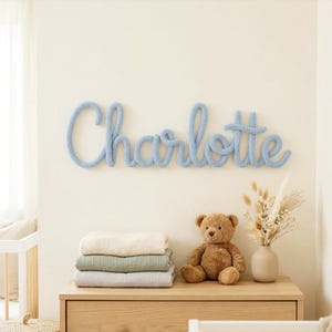 May include: A light blue knitted wall hanging spelling "Charlotte" is mounted on a cream-colored wall. Below, a wooden dresser displays folded blankets, a teddy bear, and a vase of dried flowers, creating a nursery theme.