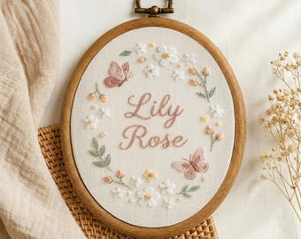 May include: Embroidered hoop art featuring the name "Lily Rose" in a cursive font, surrounded by floral and butterfly designs. The artwork is set in a wooden hoop with a metal hanging loop. The color palette includes soft pinks, whites, and greens.