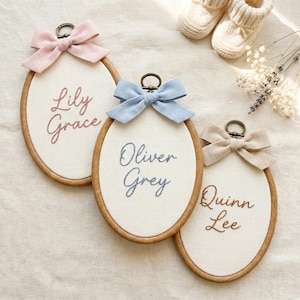 May include: Three oval wooden embroidery hoops featuring off-white fabric with embroidered names. The first hoop has a pink bow and reads Lily Grace. The second has a light blue bow and reads Oliver Grey. The third has a beige bow and reads Quinn Lee.