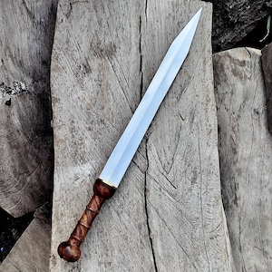 May include: A Roman gladius sword with a polished steel blade and a wooden handle. The handle is a dark brown color with a textured grip. The sword is lying on a weathered wooden surface, showcasing its historical design.