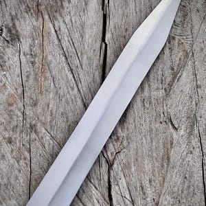 May include: A close-up of a silver sword blade with a sharp point. The blade is set against a weathered, gray wooden surface. The sword's surface reflects light, highlighting its polished finish.