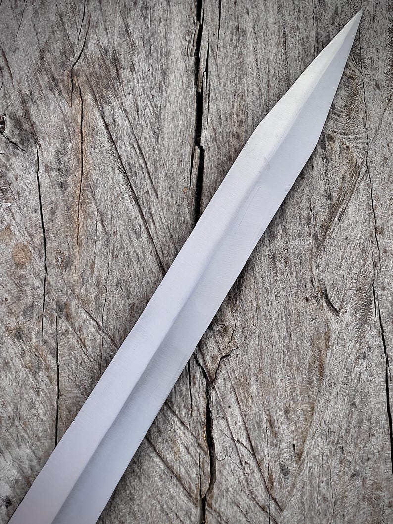 May include: A close-up of a silver sword blade with a sharp point. The blade is set against a weathered, gray wooden surface. The sword's surface reflects light, highlighting its polished finish.