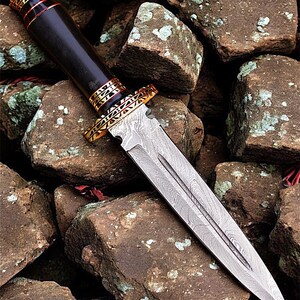 May include: A Damascus steel dagger with a dark wooden handle and gold-coloured accents. The blade has a detailed pattern, and the handle is capped with gold and red trim. The dagger rests on rough, brown stones.