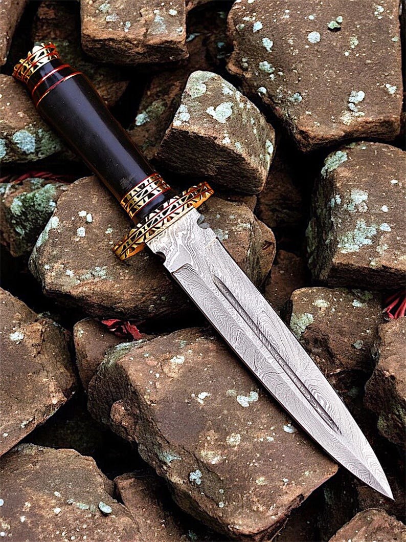 May include: A Damascus steel dagger with a dark wooden handle and gold-coloured accents. The blade has a detailed pattern, and the handle is capped with gold and red trim. The dagger rests on rough, brown stones.
