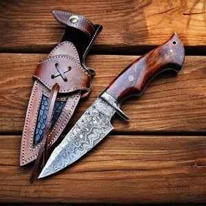 May include: A handcrafted hunting knife with a patterned steel blade and a dark wood handle. The knife rests beside a brown leather sheath with decorative stitching and a cross-shaped closure. The background is a wooden surface.