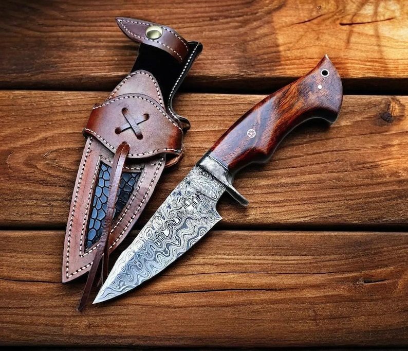 May include: A handcrafted hunting knife with a patterned steel blade and a dark wood handle. The knife rests beside a brown leather sheath with decorative stitching and a cross-shaped closure. The background is a wooden surface.