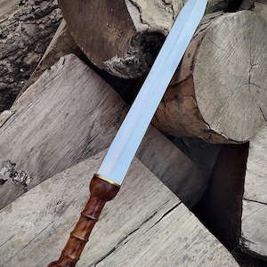 May include: A Roman gladius sword with a polished steel blade and a dark wood handle. The handle features a detailed, carved design with a brass accent near the blade. The sword is displayed against a backdrop of stacked wooden logs.