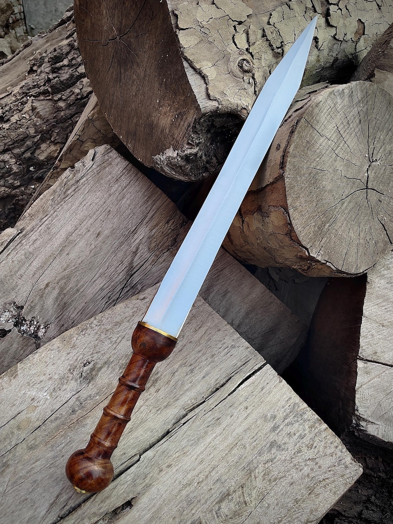 May include: A Roman gladius sword with a polished steel blade and a dark wood handle. The handle features a detailed, carved design with a brass accent near the blade. The sword is displayed against a backdrop of stacked wooden logs.