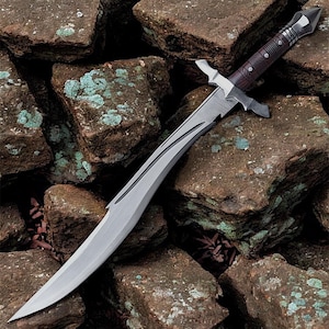 Può includere: Un pugnale argentato e marrone con una lama curva, appoggiato su un letto di rocce marroni. Il pugnale presenta un manico in legno marrone scuro con dettagli argentati e una guardia. La lama ha scanalature decorative.