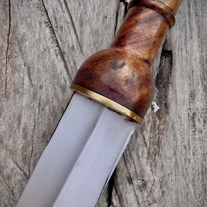 May include: Close-up of a Roman gladius sword. The handle is crafted from polished, dark wood with a brass accent at the blade's base. The blade is a polished, double-edged steel. The sword rests on a weathered wooden surface.