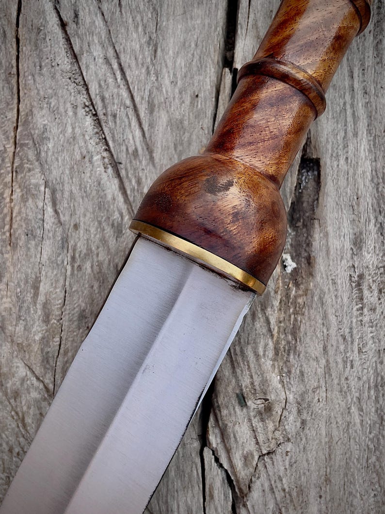 May include: Close-up of a Roman gladius sword. The handle is crafted from polished, dark wood with a brass accent at the blade's base. The blade is a polished, double-edged steel. The sword rests on a weathered wooden surface.