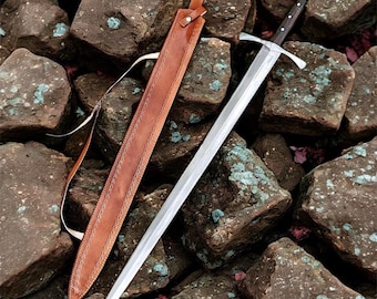 May include: A long, silver sword with a dark wooden handle and a leather sheath. The sword has a cross-shaped hilt and a pointed blade. The sheath is brown and has a strap. The sword and sheath are resting on a bed of rocks.
