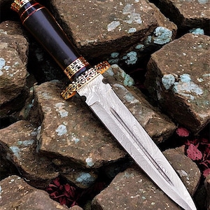 May include: A detailed close-up of a dagger with a patterned steel blade and a dark wooden handle. The handle is accented with gold-coloured metal rings and a red band. The dagger rests on a bed of rough, brown stones.