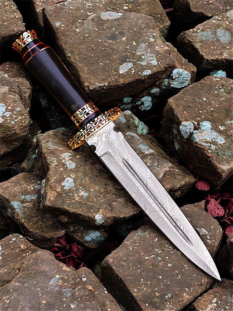 May include: A detailed close-up of a dagger with a patterned steel blade and a dark wooden handle. The handle is accented with gold-coloured metal rings and a red band. The dagger rests on a bed of rough, brown stones.
