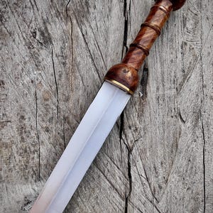 May include: A Roman gladius sword with a wooden handle and a polished steel blade. The handle is a dark brown color with a rounded pommel. The blade is straight and has a slight taper. The sword is displayed on a weathered wooden surface.