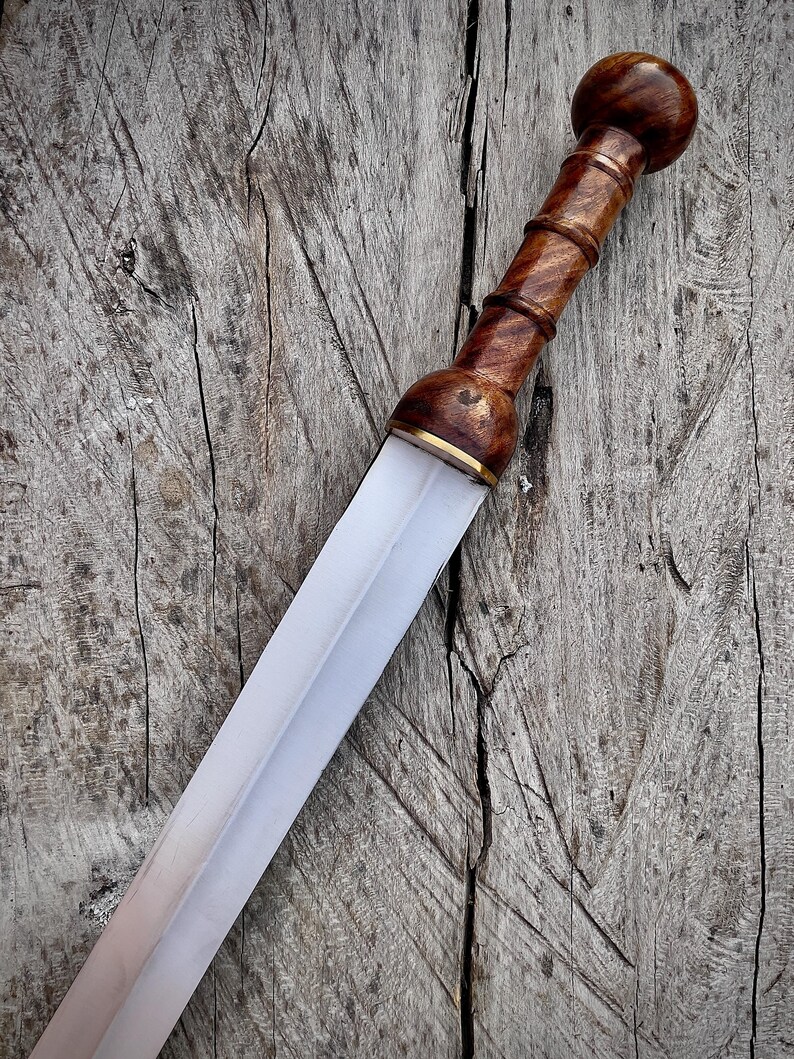 May include: A Roman gladius sword with a wooden handle and a polished steel blade. The handle is a dark brown color with a rounded pommel. The blade is straight and has a slight taper. The sword is displayed on a weathered wooden surface.