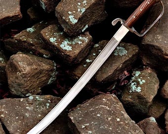 Puede incluir: Un sable con una hoja plateada curvada y un mango de madera con una guarda de metal. El mango tiene un acabado marrón oscuro con una serie de pequeños puntos de color claro. La espada se exhibe sobre un lecho de piedras marrones rugosas.
