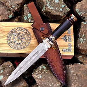 May include: A decorative dagger with a patterned blade, black handle, and gold detailing. The dagger is in a brown leather sheath, with a wooden box featuring a circular rune design. The design suggests a historical or fantasy theme.