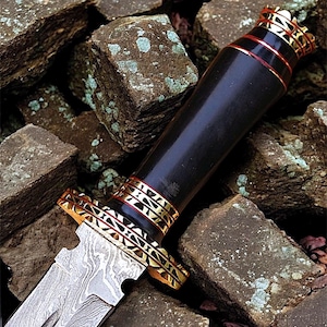 May include: A decorative dagger with a patterned blade and a black handle. The handle features gold-coloured metal and red bands. The dagger rests on a bed of rough, brown stones.