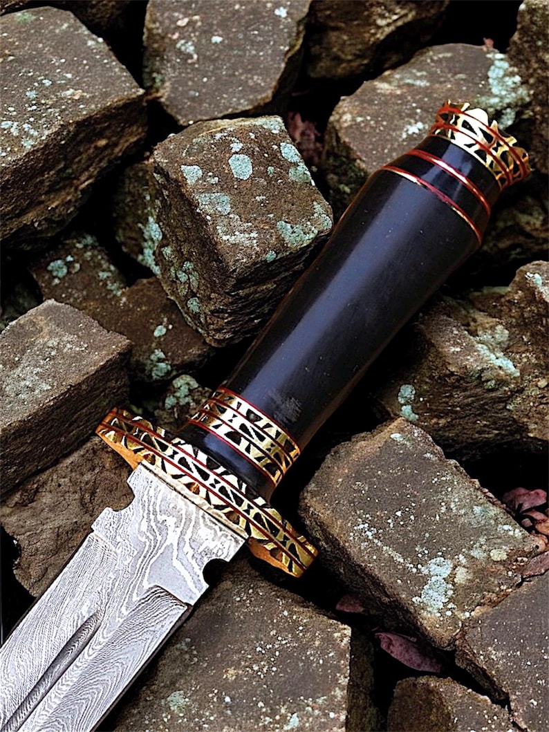 May include: A decorative dagger with a patterned blade and a black handle. The handle features gold-coloured metal and red bands. The dagger rests on a bed of rough, brown stones.