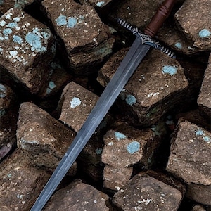 May include: A long steel sword with a dark blade and a brown wooden handle. The hilt has detailed metalwork. The sword lies on a bed of rough, brown rocks with patches of blue-green lichen.