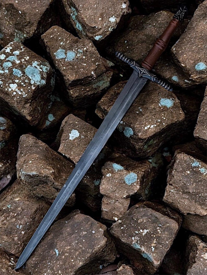 May include: A long steel sword with a dark blade and a brown wooden handle. The hilt has detailed metalwork. The sword lies on a bed of rough, brown rocks with patches of blue-green lichen.