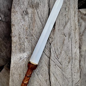 May include: A Roman gladius sword with a polished steel blade and a wooden handle. The handle is a dark brown color with a bulbous pommel and a gold-colored accent near the blade. The sword is displayed on a weathered wooden surface.