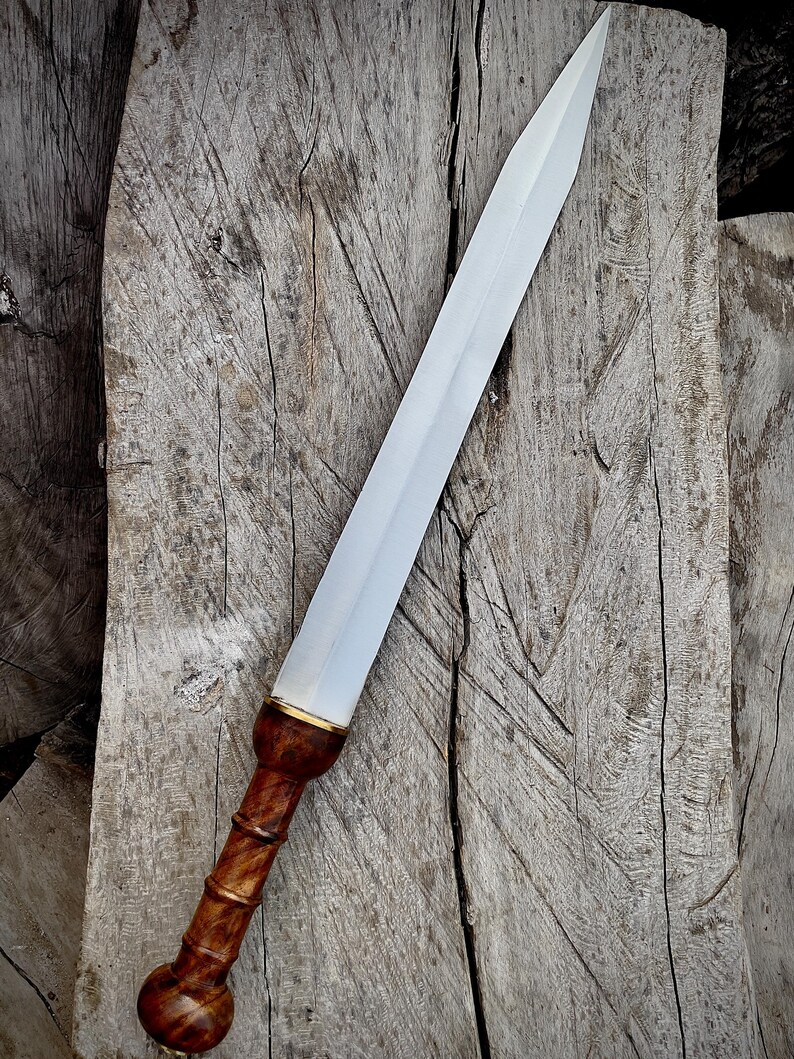 May include: A Roman gladius sword with a polished steel blade and a wooden handle. The handle is a dark brown color with a bulbous pommel and a gold-colored accent near the blade. The sword is displayed on a weathered wooden surface.