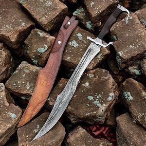 May include: A decorative sword with a patterned blade and a dark wooden handle, resting on a pile of rocks. The sword has a silver-colored guard and a curved tip. A brown leather sheath with embossed designs is next to the sword.