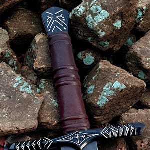 May include: A black and silver sword with a dark brown handle. The hilt and pommel have intricate white designs. The blade is dark grey, set against brown rocks. The sword's design suggests a fantasy theme.