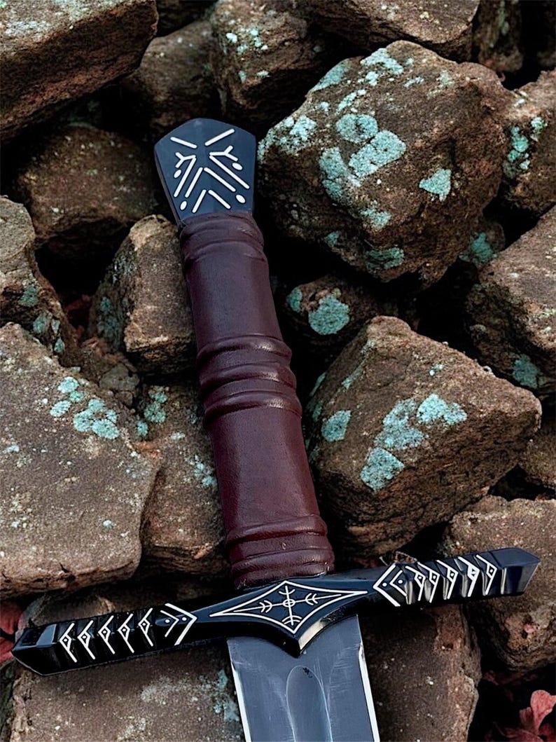May include: A black and silver sword with a dark brown handle. The hilt and pommel have intricate white designs. The blade is dark grey, set against brown rocks. The sword's design suggests a fantasy theme.