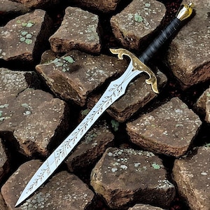 May include: A decorative sword with a silver blade featuring leaf designs. The hilt is gold-colored with ornate detailing, and the handle is wrapped in black leather. The sword rests on a bed of brown stones.