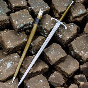 May include: A silver sword with a cross-shaped hilt and a long, pointed blade. Beside it is a green scabbard with a silver tip and a black leather band. The sword and scabbard are displayed on a bed of brown, square stones.