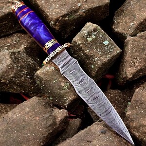 May include: A decorative dagger featuring a patterned steel blade and a purple handle. The handle has gold-coloured metal accents. The dagger is displayed on a bed of rough, brown stones.