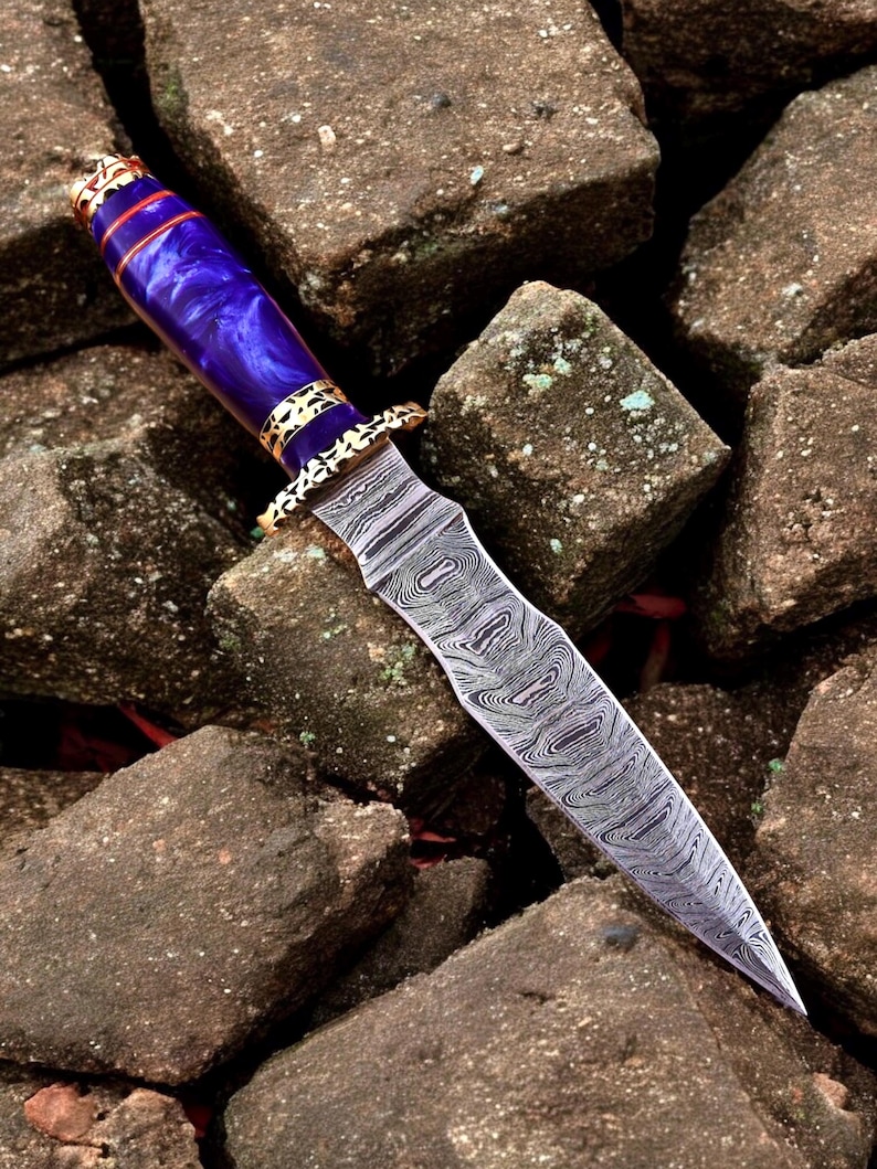 May include: A decorative dagger featuring a patterned steel blade and a purple handle. The handle has gold-coloured metal accents. The dagger is displayed on a bed of rough, brown stones.