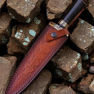 May include: A handcrafted knife with a dark wooden handle and a decorative gold-coloured metal bolster. The knife is sheathed in a brown leather case with intricate embossed patterns. The knife and sheath are set against a background of rough, brown stones.