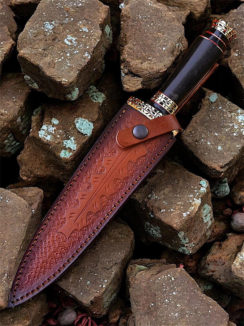 May include: A handcrafted knife with a dark wooden handle and a decorative gold-coloured metal bolster. The knife is sheathed in a brown leather case with intricate embossed patterns. The knife and sheath are set against a background of rough, brown stones.