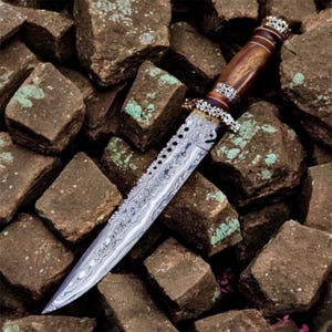 Puede incluir: Un cuchillo decorativo con una hoja de acero estampada y un mango de madera. El mango está adornado con detalles metálicos de color plateado. El cuchillo descansa sobre un lecho de piedras marrones con detalles verdes.