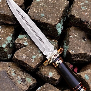 May include: A close-up of a dagger with a patterned steel blade and a dark wooden handle. The handle is accented with gold and red bands. The dagger rests on a bed of rough, brown stones.