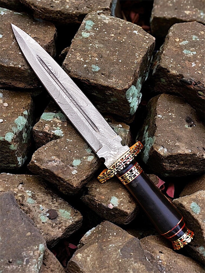 May include: A close-up of a dagger with a patterned steel blade and a dark wooden handle. The handle is accented with gold and red bands. The dagger rests on a bed of rough, brown stones.