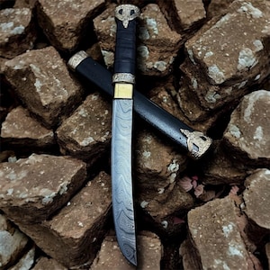 Può includere: Un coltello decorativo con una lama a fantasia e un manico nero. Il coltello è accompagnato da un fodero nero con dettagli color oro. L'impugnatura presenta un design color oro nella parte superiore.