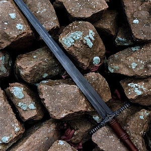 May include: A long, dark sword with a brown leather grip is positioned against a stone wall. The blade has etched markings, and the hilt features intricate designs. The sword's dark colour contrasts with the light brown stones.