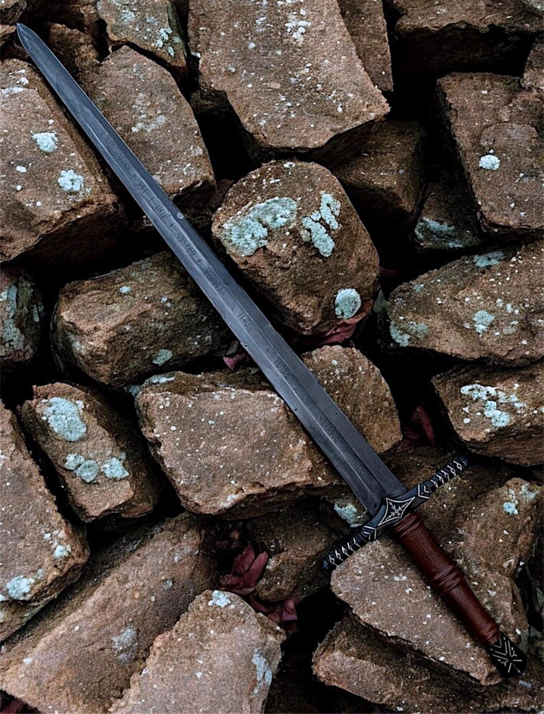 May include: A long, dark sword with a brown leather grip is positioned against a stone wall. The blade has etched markings, and the hilt features intricate designs. The sword's dark colour contrasts with the light brown stones.