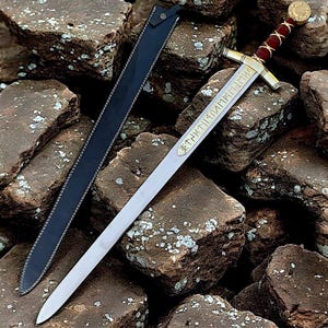 Könnte beinhalten: Ein silbernfarbenes Schwert mit goldenem Parierstange und einem rot-goldenen Griff liegt auf einem Steinhaufen. Die Klinge hat gravierte goldene Schriftzeichen. Eine schwarze Lederscheide liegt neben dem Schwert.