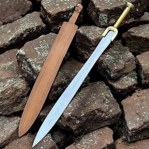 May include: A sword with a polished steel blade and a gold-colored hilt, resting on a bed of rough, reddish-brown stones. The sword is accompanied by a tan leather sheath with decorative stitching. The blade is approximately 24 inches long.