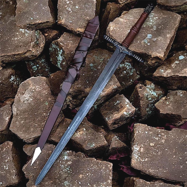 May include: A steel sword with a dark, etched blade and a brown leather-wrapped handle. The crossguard features intricate silver-coloured engravings. Beside the sword lies a matching brown leather scabbard with a silver-coloured metal tip.