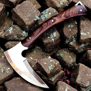 Puede incluir: Un hacha de acero pulido con una hoja curva y un mango de madera marrón oscuro. El mango está asegurado con pequeños remaches plateados. El hacha descansa sobre un montón de piedras grises rugosas.