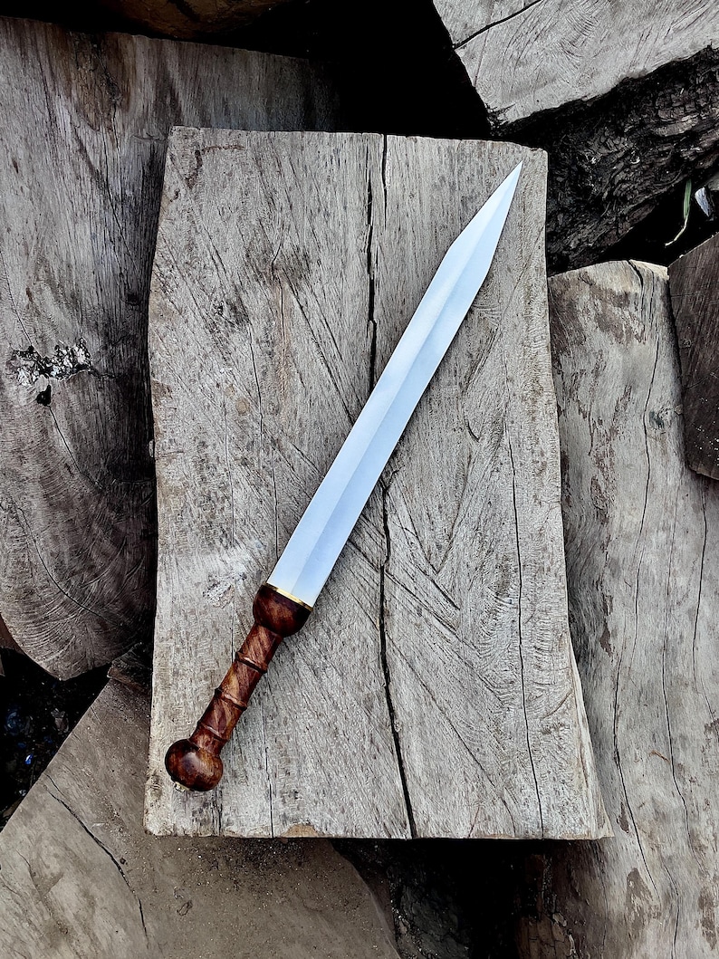 May include: A Roman gladius sword with a polished steel blade and a wooden handle. The handle is a dark brown color with a textured grip. The sword is lying on a weathered wooden surface, showcasing its historical design.