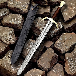 Peut inclure: Une épée décorative avec une lame à motifs, une garde transversale dorée et un manche marron. L'épée est posée sur un lit de pierres, à côté d'un fourreau en cuir noir. Le design de la lame est détaillé et les matériaux semblent de haute qualité.