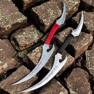 May include: Two curved daggers with silver blades and ornate handles. One dagger has a red wrapped handle, the other a black wrapped handle. The blades have an inscription. The daggers are displayed on a bed of rough, brown stones.
